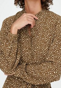 Blusa marrone a fantasia con macchie irregolari bianche, maniche lunghe con polsini a bottone e colletto alla mandarina. Tessuto morbido e leggero.
