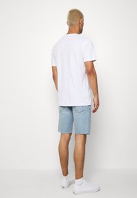 Hombre con cabello rubio corto, vistiendo una camiseta blanca básica, pantalones cortos de mezclilla azul claro, calcetines blancos y zapatillas blancas, de pie frente a un fondo blanco.