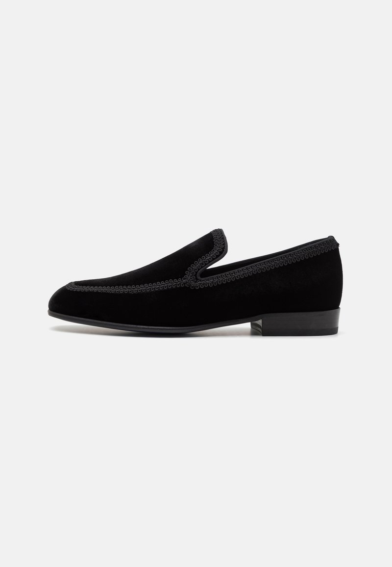Μαύρο βελούδινο παπούτσι slip-on με χαμηλό τακούνι, με διακοσμητική ραφή σε σχήμα κυματοειδούς και λεία υφή. Κλασικός σχεδιασμός με ελάχιστα αξεσουάρ.