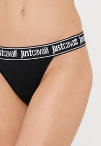 Čierne tangá nohavičky s širokým elastickým pásom s bielym nápisom "Just Cavalli". Hladký povrch a priliehavý strih.