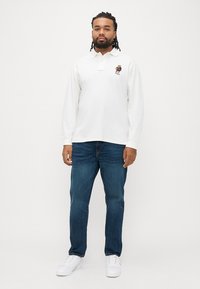 Λευκό πόλο με μακριά μανίκια και γιακά, με κεντημένο λογότυπο αρκούδας στο αριστερό στήθος. Φορεμένο με μπλε τζιν και λευκά αθλητικά παπούτσια.