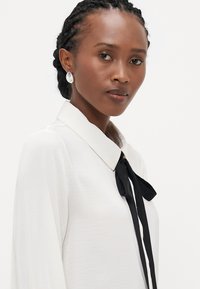 Blouse blanche avec un col classique et un nœud papillon noir. Présente des manches longues et un tissu lisse et texturé. Boucles d'oreilles rondes argentées visibles.