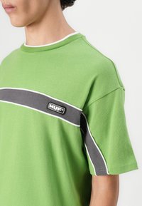 Green short-sleeve t-shirt featuring a gray horizontal stripe and white piping, with a black "HUF" logo on the chest. Cotton material.