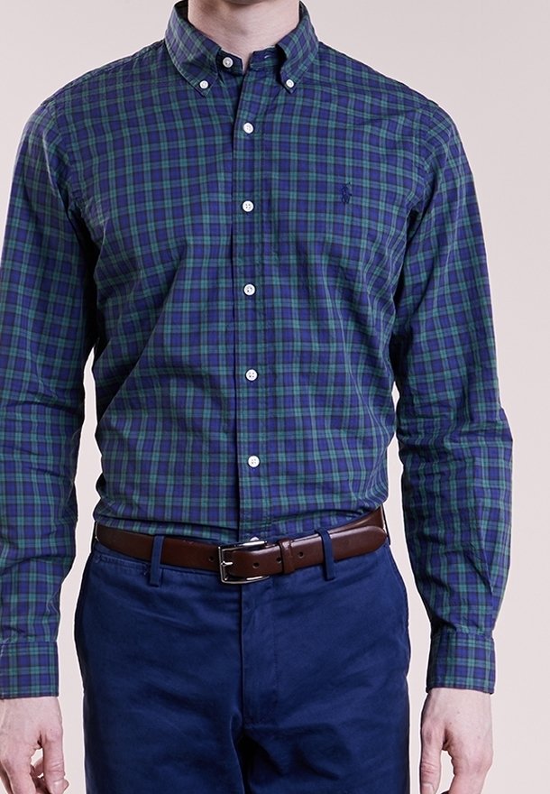 Grön och blå rutig skjorta med knappar och långa ärmar, vita knappar och en bröstlogga, i kombination med mörkblå byxor och ett brunt bälte.