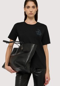 Czarny bawełniany T-shirt z haftowanym logo na piersi, w połączeniu z dużą czarną skórzaną torbą z zaokrąglonym uchwytem i błyszczącymi akcentami.