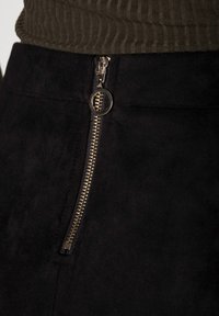 Svart mockaskirt med en synlig dragkedja och ett guldfärgat metallhandtag, placerad mot ett strukturerat olivgrönt tyg.