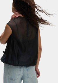 Top senza maniche nero in tessuto trasparente, leggero e dalla vestibilità ampia, con lacci sui lati. Abbinato a jeans a vita alta di colore blu chiaro.