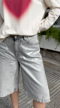 Bermudas de denim prateados com uma textura brilhante e corte regular, apresentando cinco bolsos e fecho clássico de botão. Combinadas com uma blusa clara.