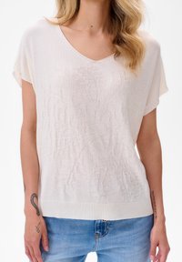 Helle beige kurzärmlige Wirkung mit V-Ausschnitt, strukturiertem Blattmuster und geripptem Saum. Kombiniert mit hellblauen Jeans.