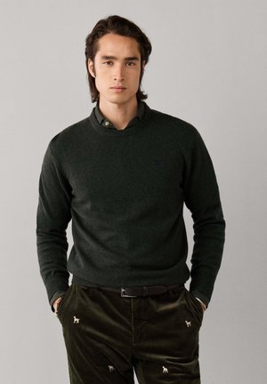 Joven con cabello oscuro lleva un suéter tejido verde oscuro sobre una camisa con cuello y pantalones de pana verde oscuro con un pequeño bordado de perro.