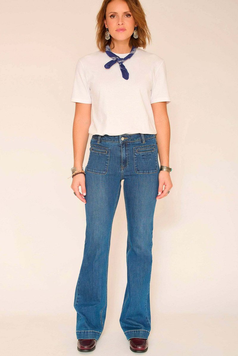 Jean bleu évasé avec une taille mi-haute et deux poches avant, associé à un t-shirt blanc et un bandana bleu autour du cou.