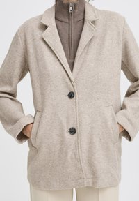 Beige wolmix jas met notch-kleppen, twee zwarte knopen, zijzakken en een relaxte pasvorm, gelaagd over een bruine rits-top.