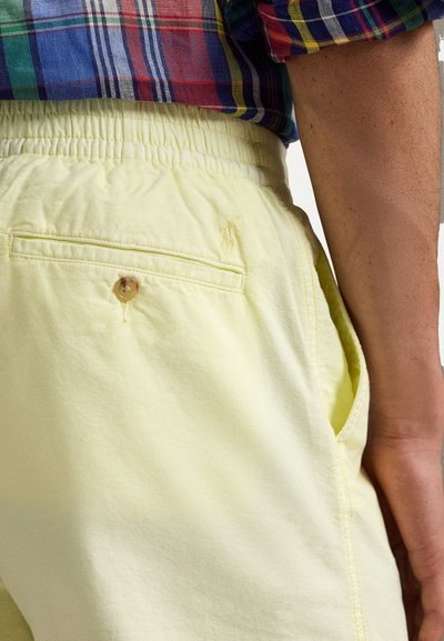 Shorts jaune clair avec une taille élastique, des poches latérales et une poche arrière avec un détail en bouton en bois. Texture de tissu lisse.
