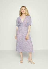Jovem mulher com cabelo louro ondulado, vestindo um vestido midi floral em lavanda com mangas bufantes e sandálias de salto brancas com tiras, posando contra um fundo simples.