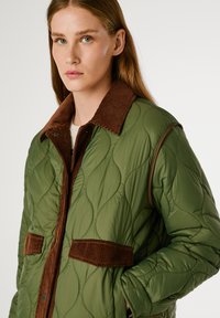 Giacca verde trapuntata con colletto e dettagli in velluto a coste marrone. Presenta una texture liscia e due tasche orizzontali nella parte anteriore.