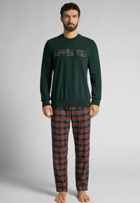 Sweatshirt en polaire verte avec "APRES SKI" en lettres à carreaux, associé à un pantalon de pyjama à carreaux rouge et marine. Texture douce, coupe décontractée.