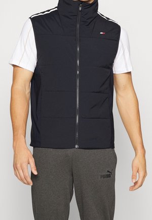 Hombre con chaleco sin mangas negro con cremallera sobre camisa blanca de manga corta y pantalones deportivos grises Puma, de pie sobre fondo liso.