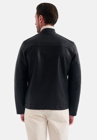 Zwarte leren jas met een gestructureerd ontwerp, met een hoge kraag en naaddetails, gedragen over lichtgekleurde broeken. Zachte textuur.