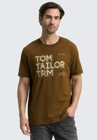 Brun bomulds T-shirt med fed grafisk tekst "TOM TAILOR" og "EMBRACE THE WILD" i lyse farver, kortærmede og klassisk rund halsudskæring.