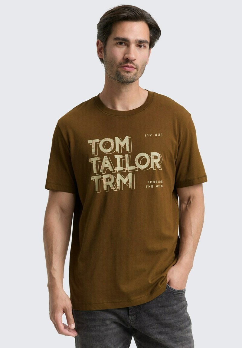 Brun bomulds T-shirt med fed grafisk tekst "TOM TAILOR" og "EMBRACE THE WILD" i lyse farver, kortærmede og klassisk rund halsudskæring.