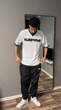 Νεαρός άνδρας που προσαρμόζει λευκό αθλητικό πουκάμισο Kappa, φοράει μαύρο παντελόνι Kappa και λευκά αθλητικά παπούτσια, στέκεται μπροστά σε καθρέφτη ολόσωμο μέσα σε εσωτερικό χώρο.