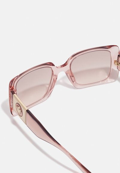 Óculos de sol oversized em rosa, com uma armação transparente e lentes degradê. Os detalhes em dourado nas hastes têm um detalhe em forma de logotipo redondo.