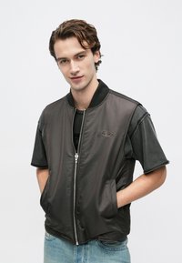 Schwarze Bomberjacke mit Reißverschluss, geripptem Kragen und dezentem Logodetail. Darüber getragen ein graues Kurzarmshirt, kombiniert mit hellen Jeans.