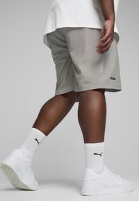 Pantalones cortos gris claro de tela ligera con un bolsillo lateral con cremallera, combinados con zapatillas blancas y calcetines blancos de canalé que llevan un logo negro.