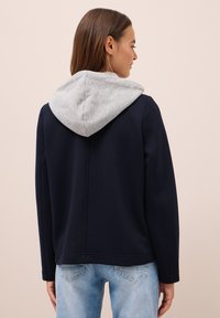 Femme avec des cheveux bruns raides portant une veste marine avec une capuche grise et un jean bleu clair, se tenant devant un fond beige.