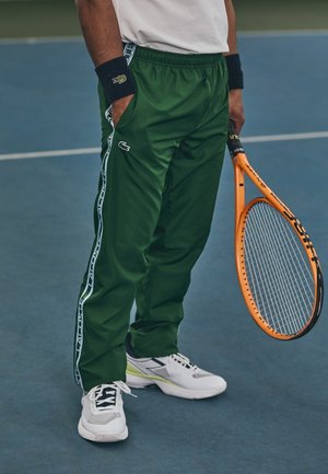 Person trägt grüne Lacoste Trainingshose, weiße Sneakers, schwarze Handgelenkbänder und hält einen orangefarbenen Tennisschläger auf einem blauen Platz.