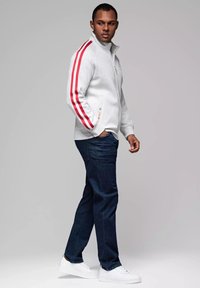 Giacca grigia con zip e strisce rosse sulle maniche, abbinata a jeans blu scuro e sneakers bianche. Il tessuto appare liscio e casual.