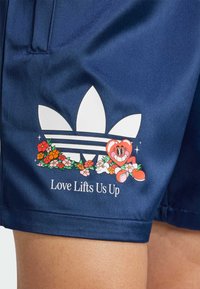 Marinblå shorts i slätt tyg med en vit Adidas trefoil-logotyp som har blommor och en tecknad karaktär, pryds av texten: "Kärlek lyfter oss upp."