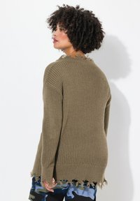 Pull en mailles vert olive à coupe ample, au tissu texturé et avec des détails de bord effiloché. Les manches longues dépassent le poignet.