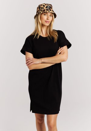 Vrouw draagt een zwart jurkje met korte mouwen en een emmerhoed met luipaardprint, staand met gekruiste armen tegen een effen achtergrond.