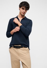 Marineblå langærmet polo med krave, med en halv lynlås. Kombineret med beige bukser og et sort bælte. Glat stoftekstur.