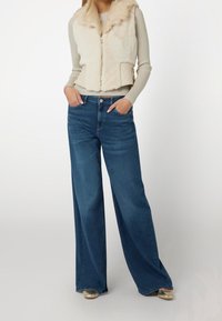 Veste en fausse fourrure crème avec côtés matelassés, associée à un jean bleu foncé à jambes larges, avec une taille haute et des poches avant. Chaussures dorées visibles.