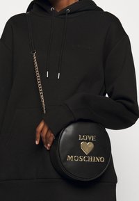 Borsa a tracolla rotonda nera in pelle con lettering dorato embossato "LOVE" e un cuore, dotata di una tracolla a catena dorata. Superficie testurizzata.