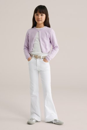 Jauna meitene stāv ar rokām kabatās, valkājot lillā kardiganu, baltu topu, baltas izplūdušas bikses, baltu jostu un sudrabotas sporta kurpes uz neitrāla fona.