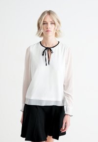 Blouse blanche avec des manches longues transparentes, un col à bord noir, et une fermeture à nouer au niveau du cou. Présente une texture lisse et une coupe légèrement ample.