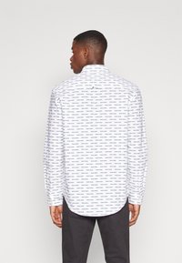 Camicia bianca a maniche lunghe con un motivo di testo ripetuto, dotata di polsini con bottone e orlo curvo. La vista posteriore mostra un piccolo etichetta con logo.