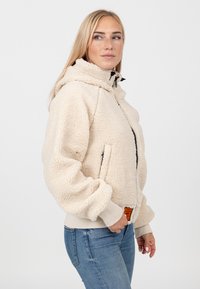 Kremowa polarowa kurtka z zamkiem z przodu, dużym kapturem i elastycznymi mankietami. Posiada boczne kieszenie oraz pomarańczową metkę marki przy dolnym brzegu.