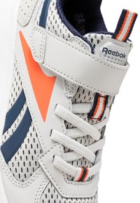 Baskets blanches et grises avec un matériau en maille, accents orange et marine, bande Velcro et surface texturée. Comprend des lacets et un détail de logo.