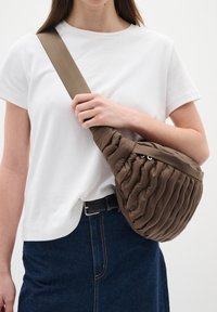 Borsa a spalla marrone trapuntata di forma arrotondata, con una superficie testurizzata e una tracolla beige regolabile, indossata su una camicia bianca.