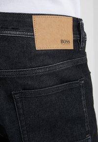 Detailní záběr na zadní kapsu černých džín s hnědým koženým štítkem s raženým nápisem "BOSS HUGO BOSS" na pase.