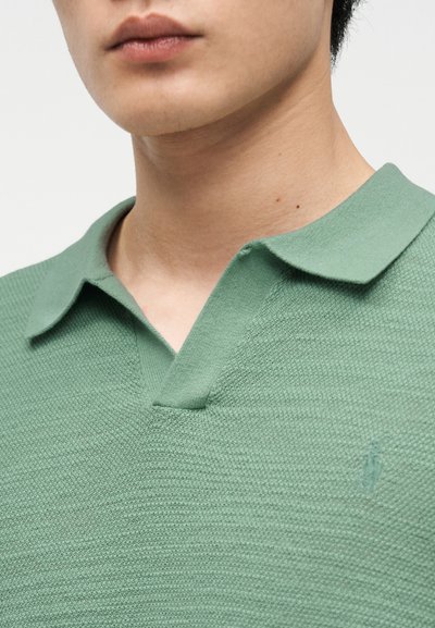 Κοντινό πλάνο ενός ατόμου που φοράει πράσινο πόλο πουκάμισο με υφή και γιακά, εμφανίζοντας μόνο τον λαιμό και το κάτω μέρος του προσώπου.