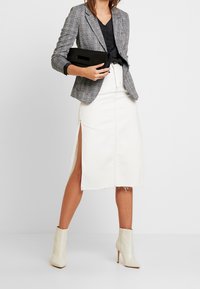 Grijze geruite blazer over een zwarte blouse, gecombineerd met een witte knielange rok met zijslits, en een crèmekleurige enkellaarsjes. Zwarte clutch.
