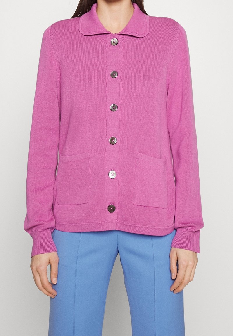 Femme portant un cardigan rose boutonné avec deux poches devant et un pantalon bleu, debout devant un fond blanc uni.