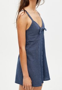 Robe en denim à rayures bleu foncé avec un décolleté en V et des bretelles réglables. Présente un nœud à l'avant et une coupe évasée et ample.
