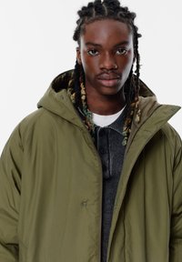 Olivengrøn hættejakke med en glat tekstur, prydet med trykknapper, lagdelt over en mørkegrå skjorte med knapper.