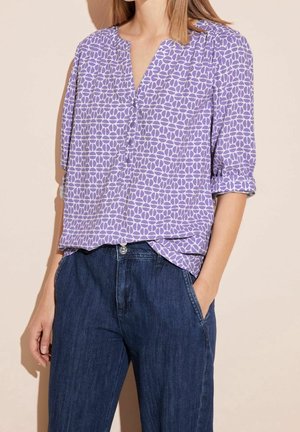 Vrouw die een paarse blouse met patroon draagt, met opgerolde mouwen, ingestopt in een high-waisted donkerblauwe spijkerbroek, met haar handen in de zakken.